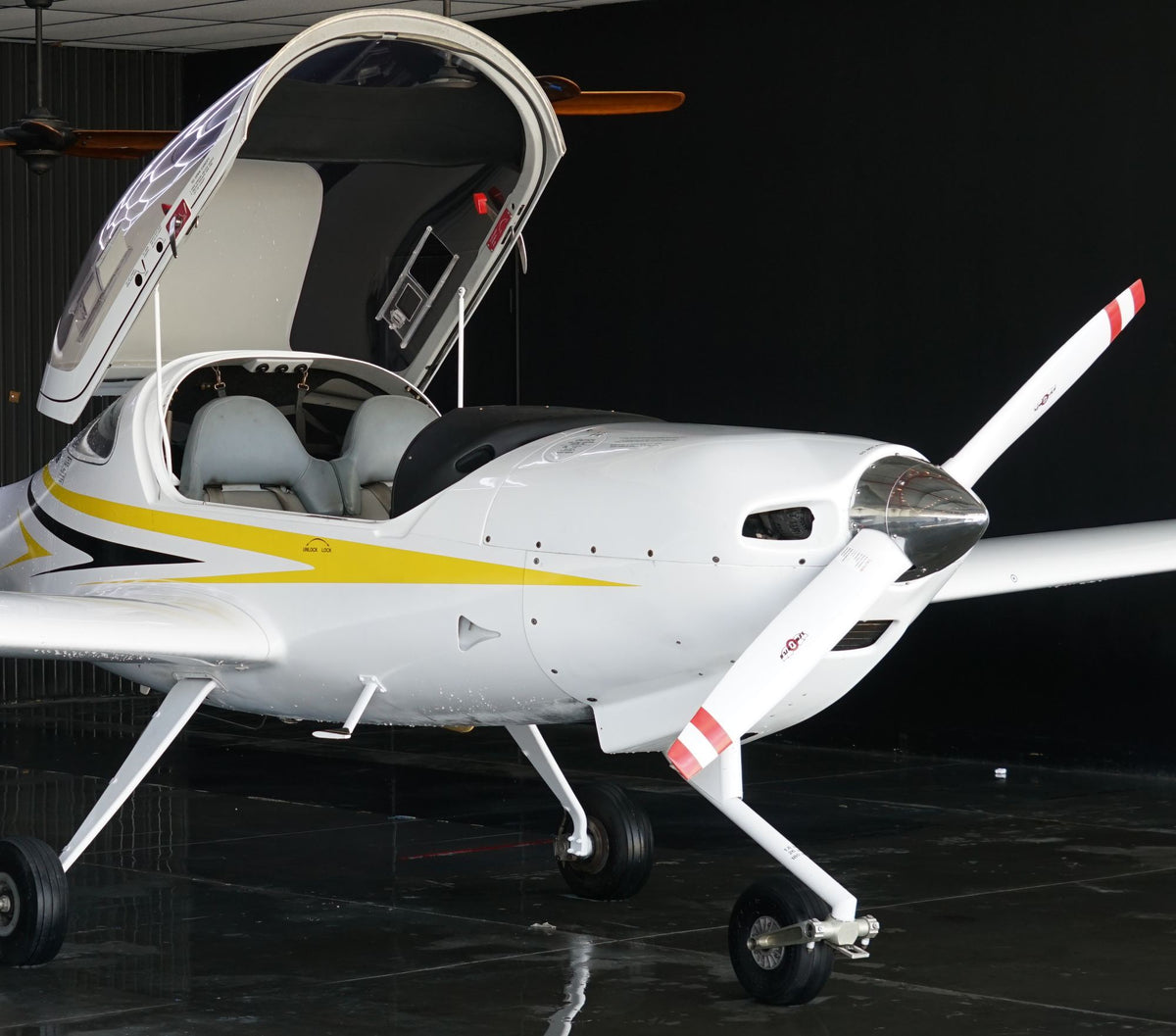 White Diamond DA20 trainer with yellow stripes and its canopy raised, parked in a dark hangar