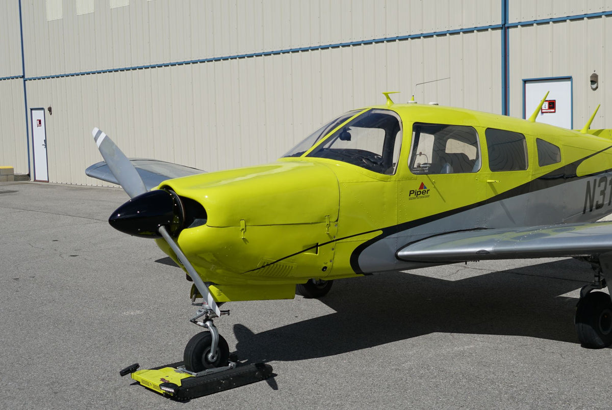Front-quarter view of a yellow Piper Arrow parked by a tan hangar wall in bright sun