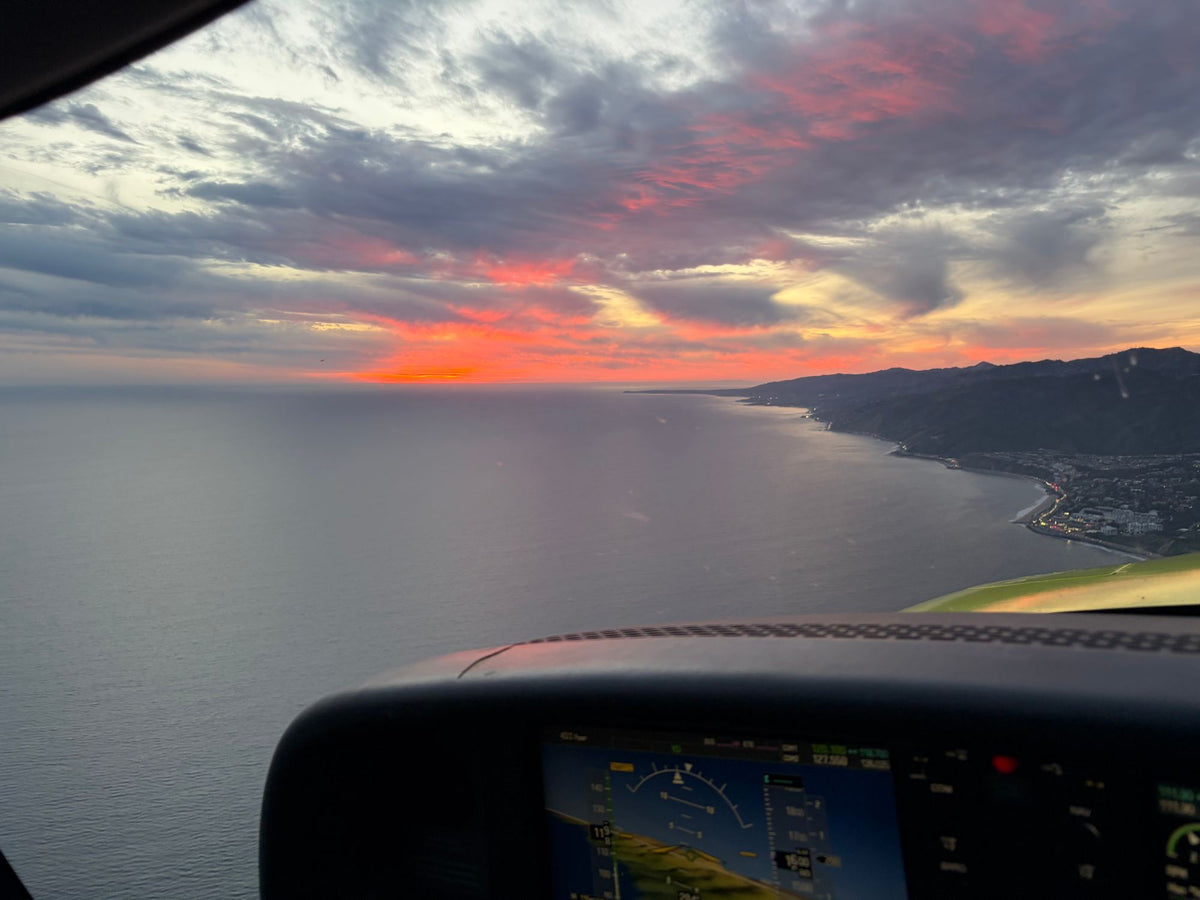 View over an aircraft instrument panel of the coastline and hills curving along the ocean under a red sunset sky