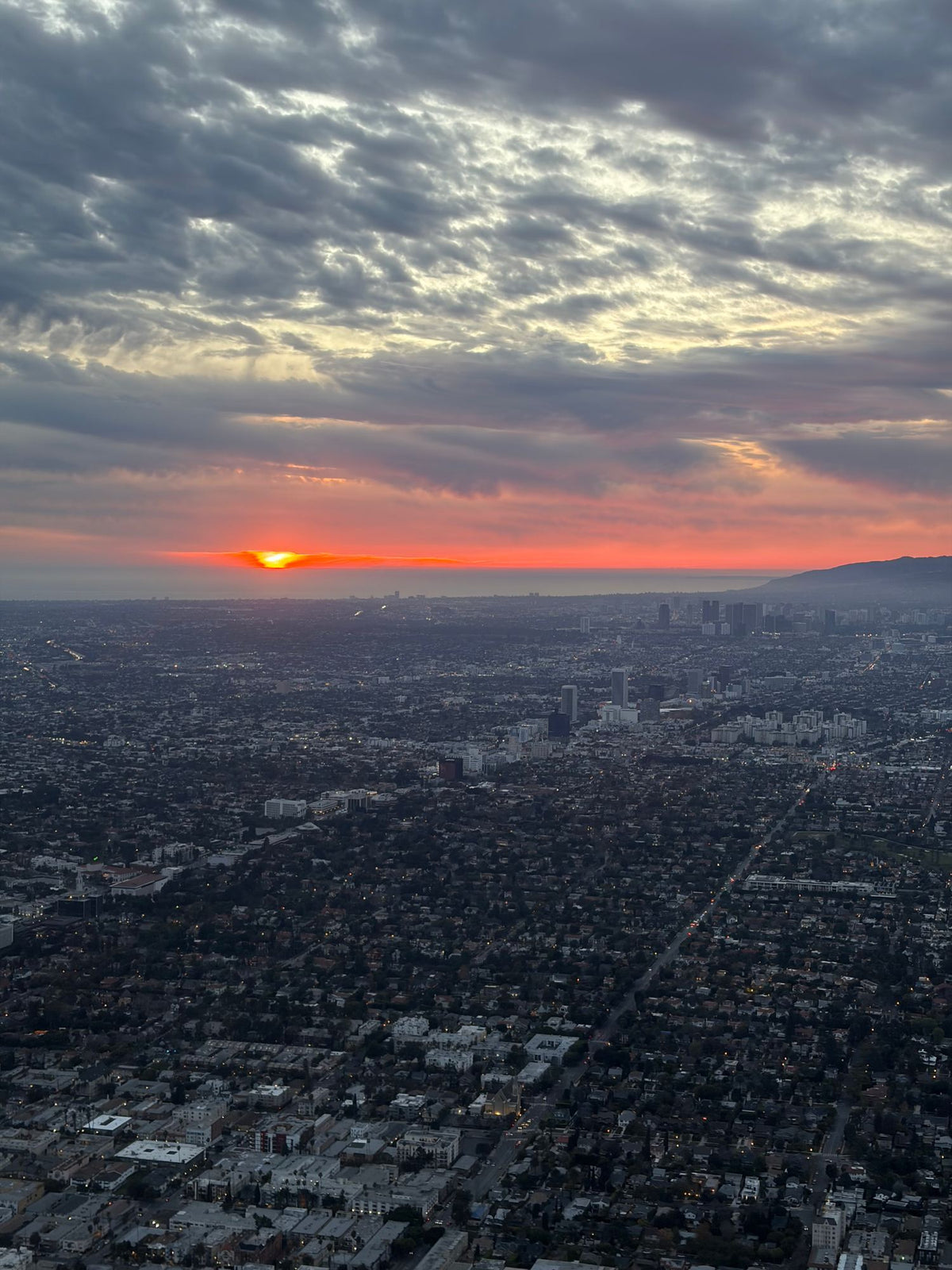Aerial view at sunset across Los Angeles neighborhoods toward the ocean, the sun on the horizon under a cloudy sky