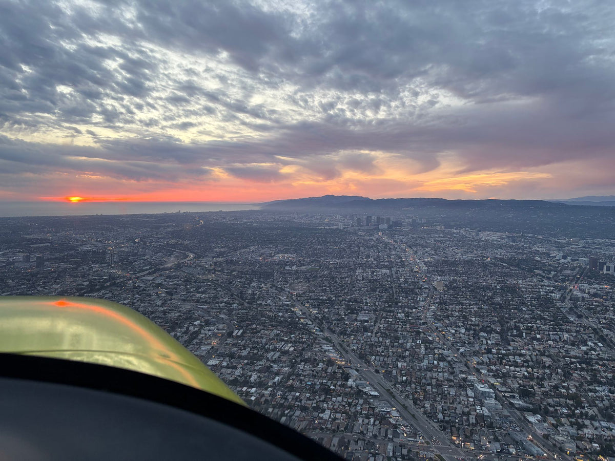 Yellow aircraft wing in the corner of the frame above Los Angeles streets at dusk, sun setting over the ocean