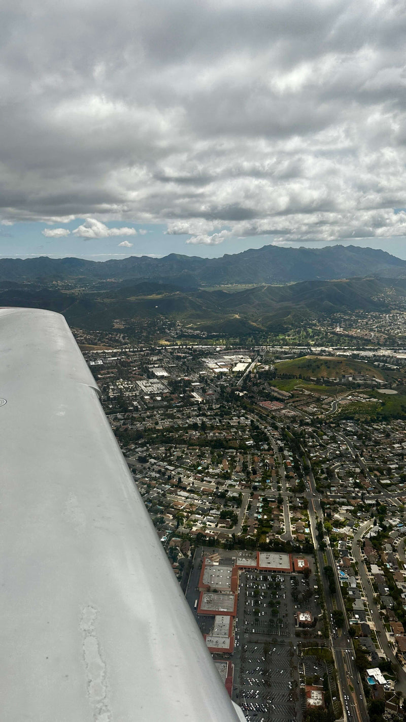 The wing of a small airplane over the San Fernando Valley on the way back to Van Nuys, with the city grid below and mountains under the clouds on the horizon