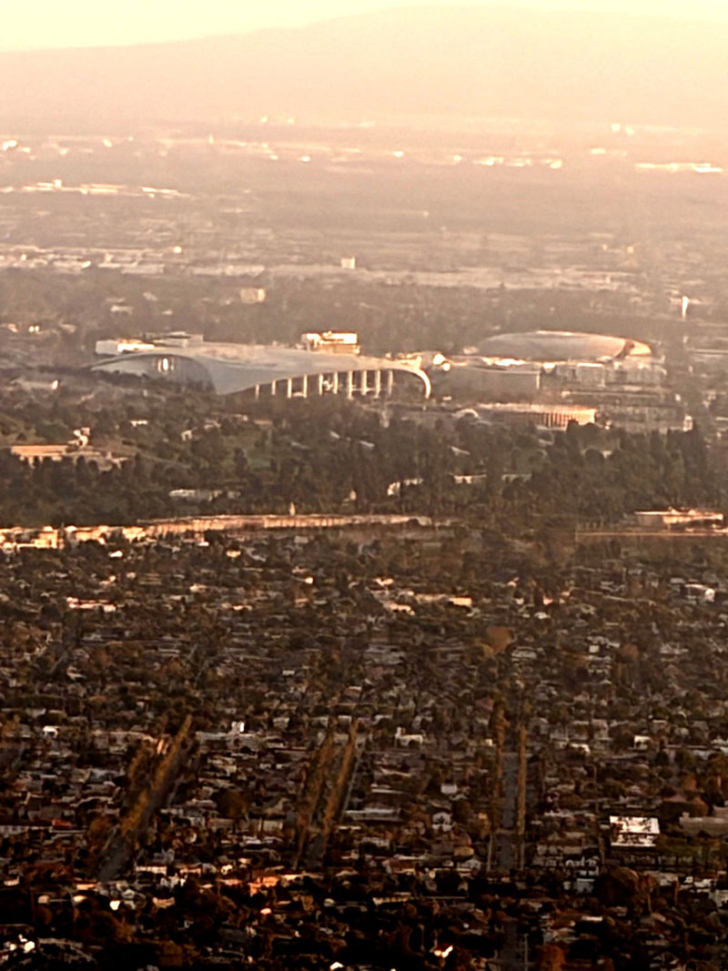 SoFi Stadium in Inglewood at golden hour, seen from a small airplane passing just north of it, with the neighborhoods of South Los Angeles in the foreground